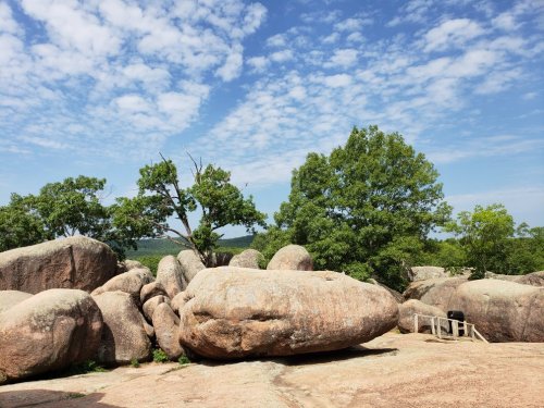 A rocky landscape with large boulders and green trees under a blue sky with scattered clouds.