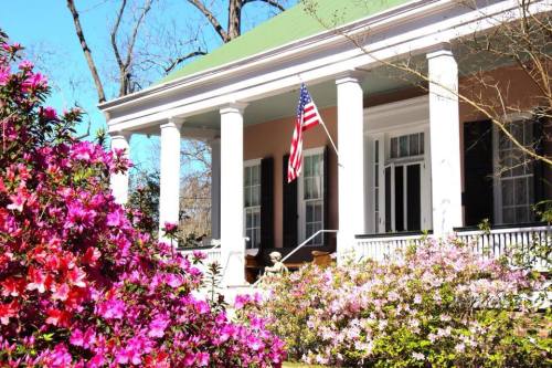 A charming house with a green roof, white columns, and an American flag, surrounded by vibrant pink flowers.