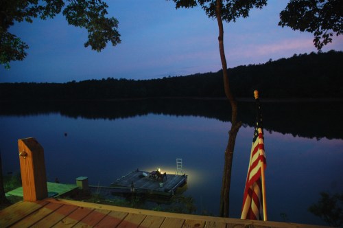 A serene lake at dusk, with a dock illuminated by a light and an American flag gently waving nearby.