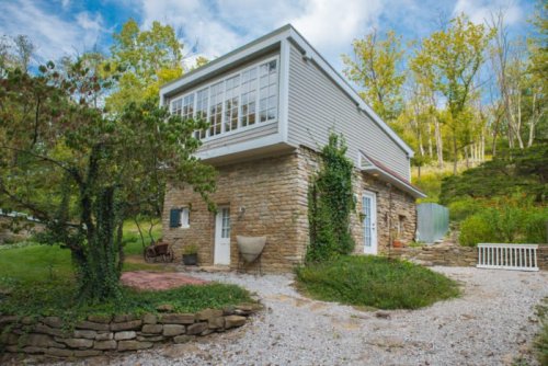 A modern two-story house with stone and wood exterior, surrounded by greenery and a gravel pathway.