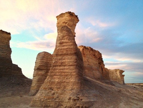 A tall, weathered rock formation stands against a colorful sky at dusk, surrounded by arid landscape.
