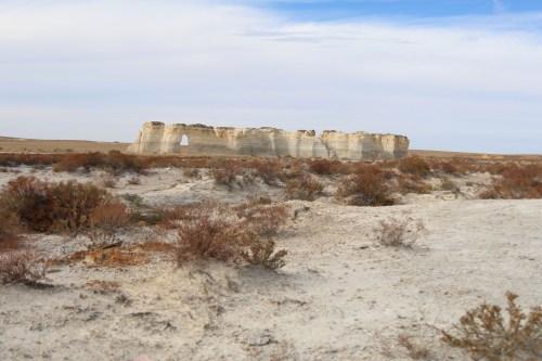 A rocky formation stands in a barren landscape, with sparse vegetation and a cloudy sky in the background.