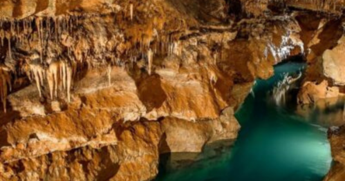 A serene underground cave with orange rock formations and a turquoise water stream flowing through.