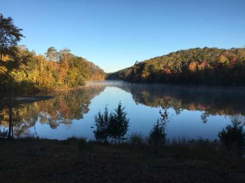 A serene lake surrounded by autumn foliage, reflecting trees and a clear blue sky. Mist rises from the water's surface.