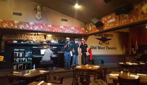 A stage in an Old West theater with performers and a pianist, surrounded by rustic decor and empty dining tables.