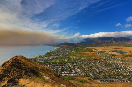 A panoramic view of a coastal town with mountains and clouds, showcasing a mix of land and sea under a blue sky.