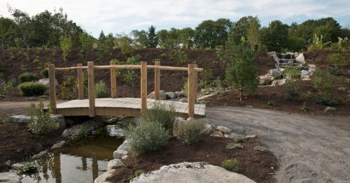 A wooden bridge over a small pond in a landscaped garden with trees and rocks, featuring a gentle waterfall in the background.