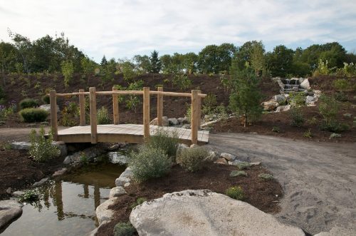 A wooden bridge over a small stream in a landscaped garden with rocks and greenery under a cloudy sky.