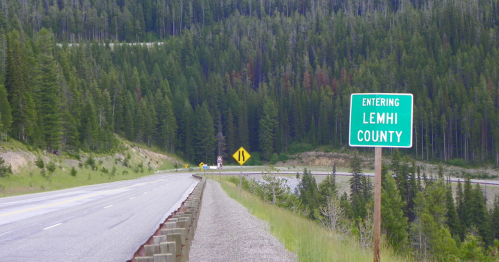 A green sign reading "Entering Lemhi County" beside a winding road surrounded by dense forested mountains.