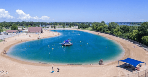 Aerial view of a sandy beach area with a blue lagoon, water activities, and trees under a clear blue sky.