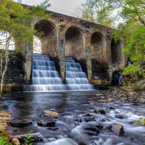 A stone bridge with arches over a river, featuring two waterfalls cascading into a rocky stream below.