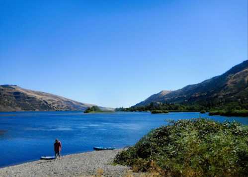 A person stands by a calm river surrounded by hills and greenery under a clear blue sky.