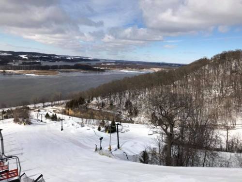 Snow-covered landscape with a river and hills under a partly cloudy sky. Ski lift visible in the foreground.