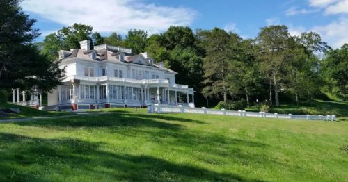 A large, elegant white mansion surrounded by lush green grass and trees under a clear blue sky.