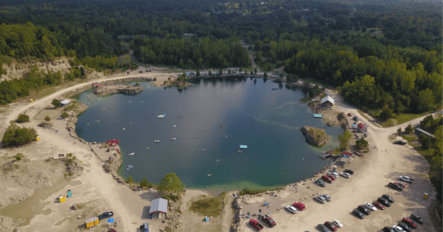 Aerial view of a serene lake surrounded by trees, with people swimming and parked cars along the shore.