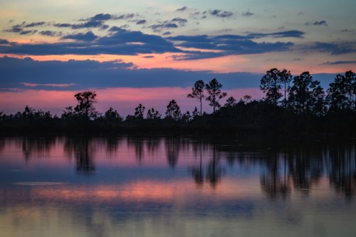 A serene sunset over a calm lake, with colorful reflections and silhouetted trees along the shoreline.