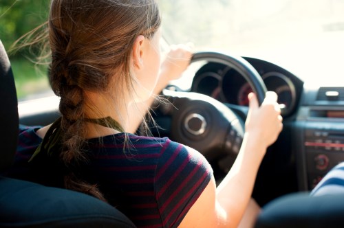 A person with a braid drives a car, focusing on the road ahead. Sunlight filters through the windshield.