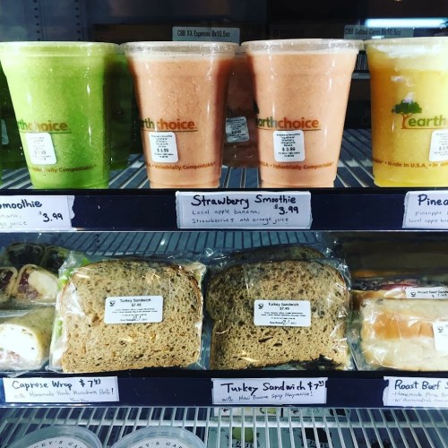 Four smoothie cups in various colors and sandwiches displayed on a shelf, with prices labeled underneath each item.
