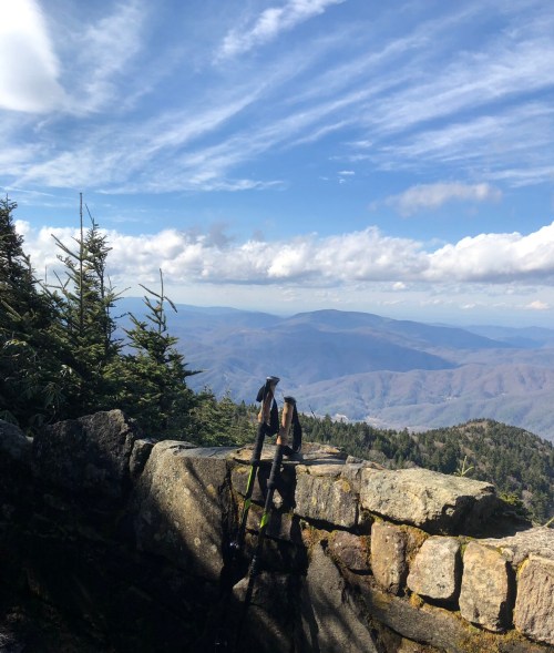 Hiking poles rest on a rocky ledge, overlooking a vast mountain landscape under a blue sky with scattered clouds.