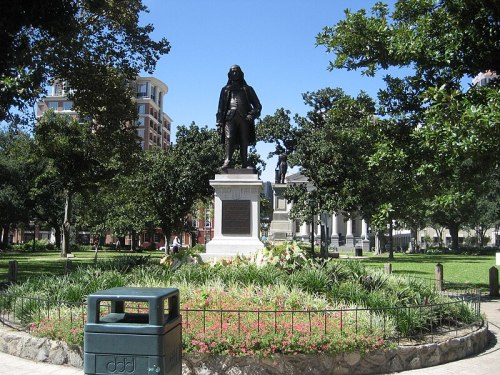 A statue of a historical figure stands in a park surrounded by greenery and flowers, with buildings in the background.