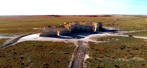 Aerial view of a large rock formation in a vast, open landscape with sparse vegetation and a clear blue sky.