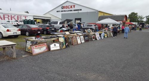 A flea market scene with vendors displaying various items and people browsing near a large building.