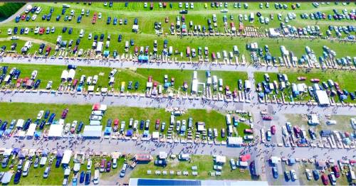 Aerial view of a large outdoor event with rows of cars, vendors, and crowds on green grass.