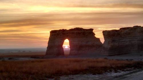 Sunset through a natural rock arch, with vibrant orange and yellow hues illuminating the sky and surrounding landscape.