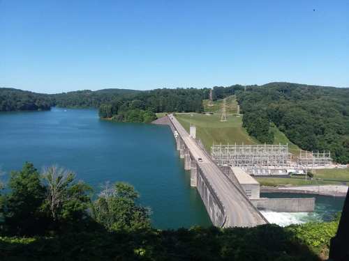 A scenic view of a dam with water flowing, surrounded by lush green hills and a clear blue sky.