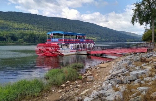 A riverboat with red paddle wheels docked by a rocky shore, surrounded by green hills and a partly cloudy sky.