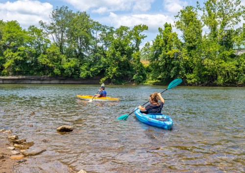 Two people kayaking on a calm river surrounded by lush greenery and a partly cloudy sky.