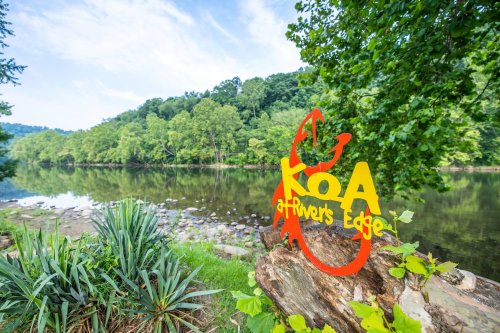 Colorful KOA sign at River's Edge by a serene river surrounded by lush greenery and trees.