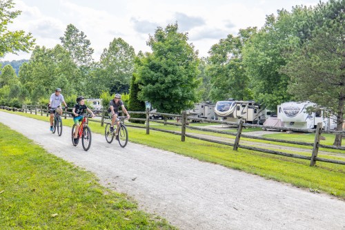 Three people ride bicycles along a gravel path near RVs and lush greenery in a campground setting.