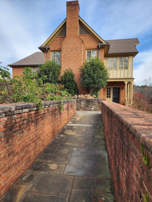 A brick pathway leads to a large, two-story house with a chimney and greenery on either side.