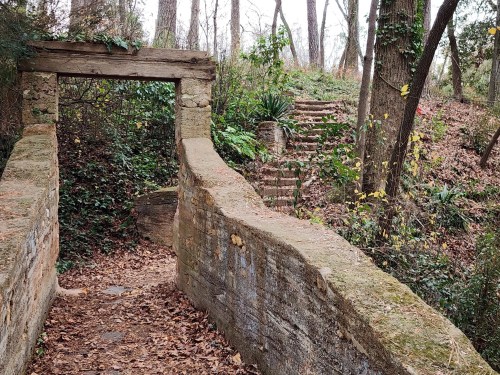 A stone archway leads to a path with steps, surrounded by trees and fallen leaves in a serene forest setting.