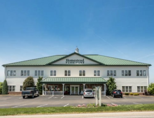 A large furniture store with a green metal roof and white exterior, featuring a parking lot and clear blue sky.