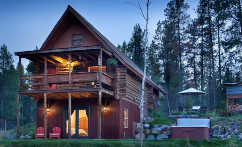 A cozy wooden cabin with a balcony, surrounded by trees, featuring a hot tub and outdoor seating.