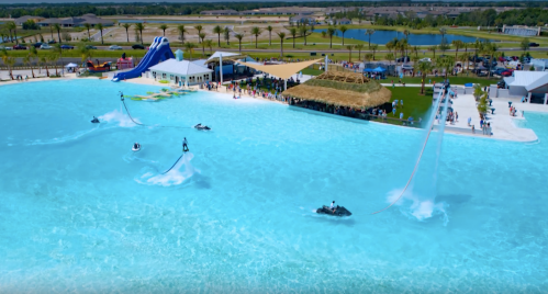 Aerial view of a water park with jet skis and a large pool, featuring palm trees and a crowd enjoying the sunny day.