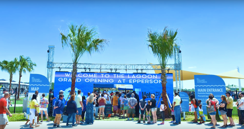 A crowd gathers at the Lagoon grand opening event, with palm trees and a blue sky in the background.