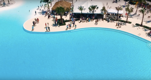Aerial view of a vibrant blue pool area with people enjoying the sun and palm trees in a tropical setting.