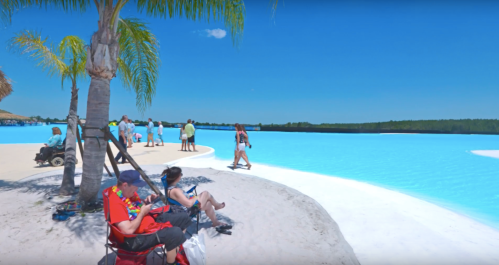 A sunny beach scene with people relaxing, walking, and enjoying a vibrant blue lagoon under a clear sky.