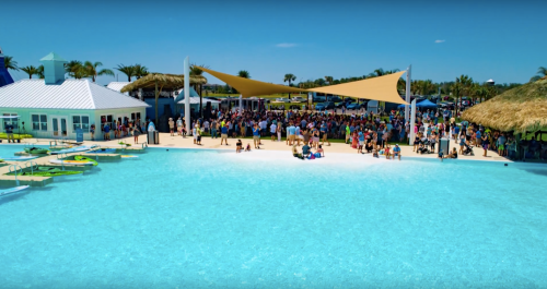 A vibrant beach scene with a large crowd gathered near a pool and shaded areas, surrounded by palm trees and blue skies.