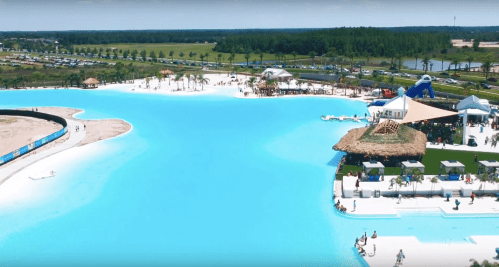 Aerial view of a vibrant blue lagoon with sandy shores, palm trees, and recreational areas filled with people.