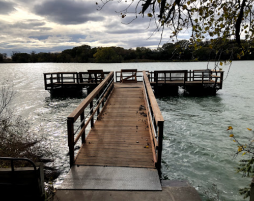 A wooden dock extends into a calm lake, surrounded by trees under a cloudy sky.