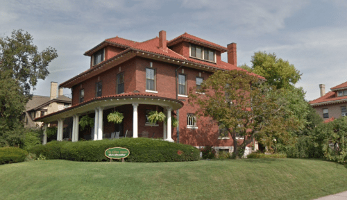 A large red brick house with a porch, surrounded by greenery and trees, under a cloudy sky.