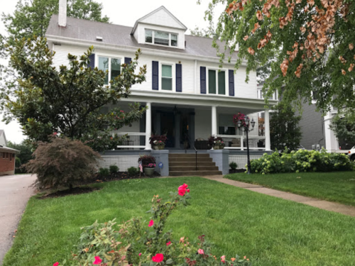 A large white house with blue shutters, a front porch, and landscaped yard featuring flowers and greenery.