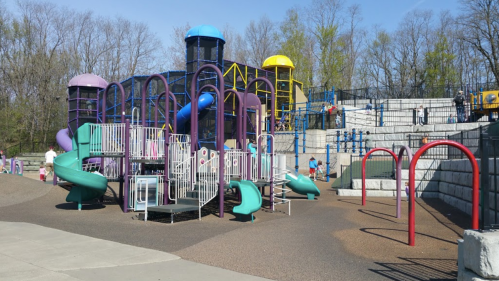 Colorful playground with slides and climbing structures, surrounded by trees and a gravel area. Sunny day.