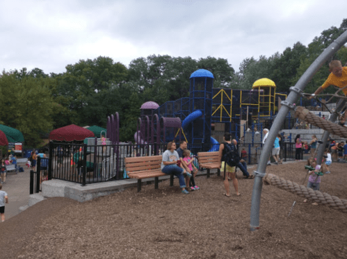A playground with colorful structures, benches, and children playing, surrounded by trees under a cloudy sky.