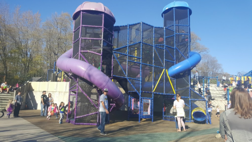 Colorful playground structure with slides and climbing areas, surrounded by children and adults enjoying the day.