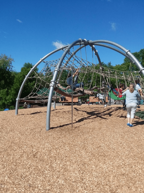 A playground with a large climbing structure and children playing on wood chip ground under a clear blue sky.